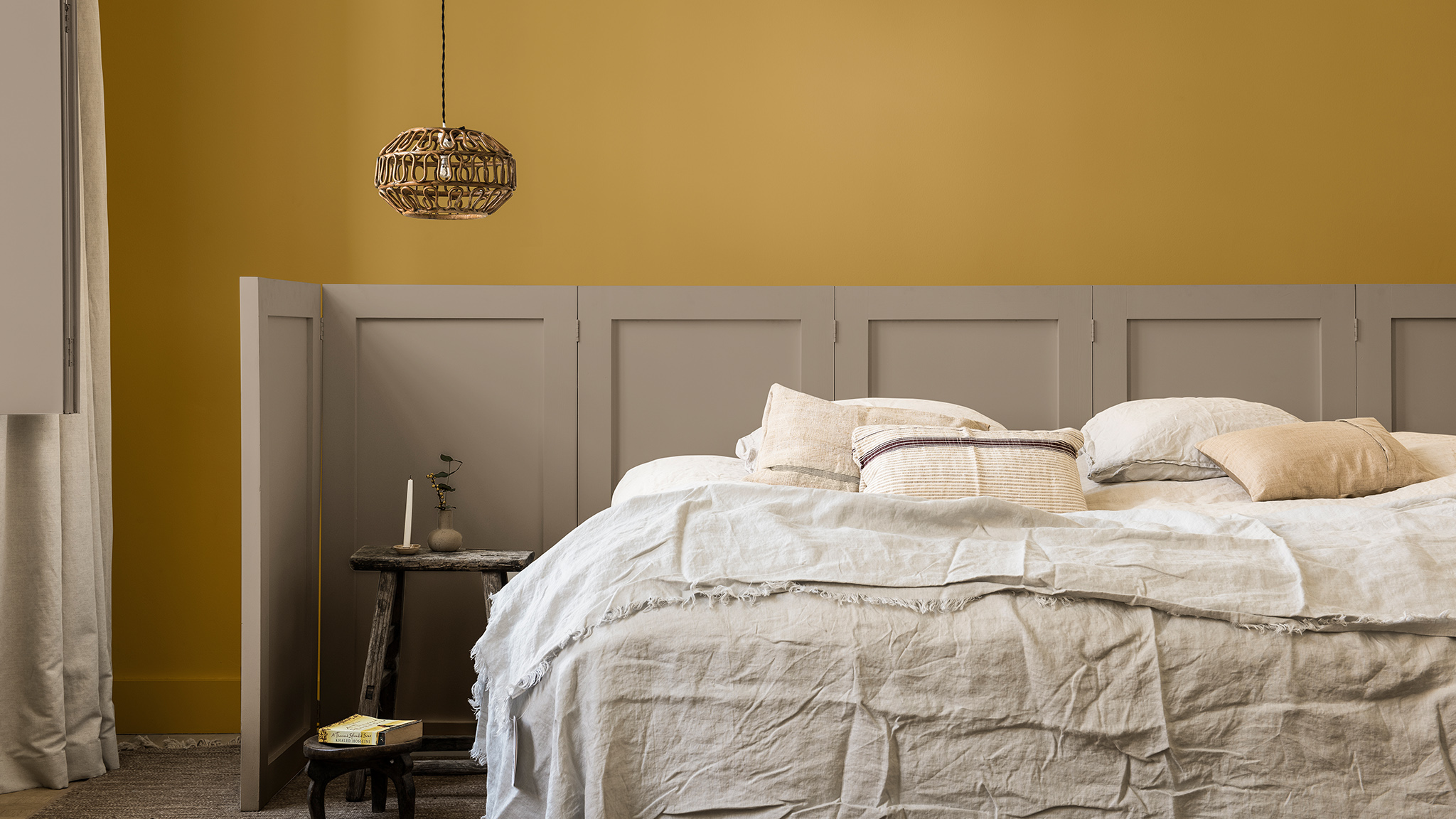 Slaapkamermuur in een goudgeel en Brave Ground geschilderd, met een houten scherm achter het bed Slaapkamermuur in een goudgeel en Brave Ground geschilderd, met een houten scherm achter het bed